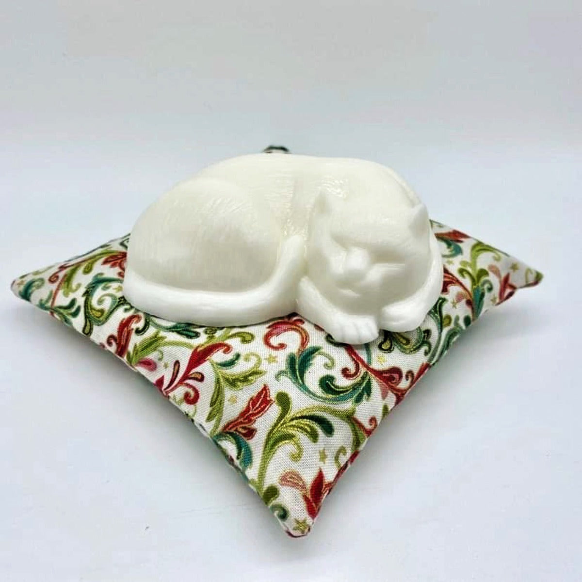 White sleeping cat-shaped soap on a red, green, gold and white paisley print square pillow