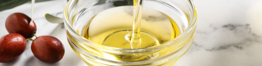 Jojoba oil in a clear glass bowls with three jojoba seeds on a white marble background