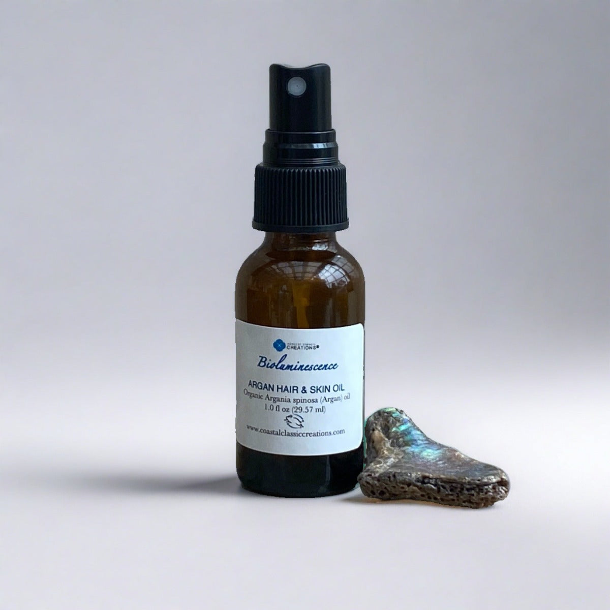 Bioluminescence Argan Hair & Skin Oil bottle with a spray top, alongside a small abalone shell
