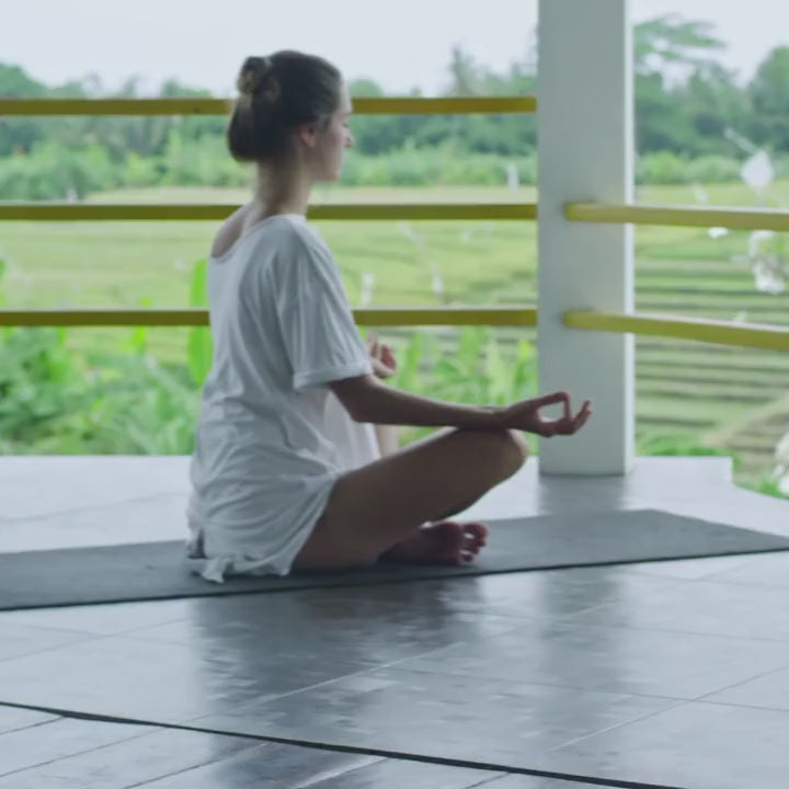 Relaxing meditation video featuring a woman practicing seated meditation on a yoga mat in a peaceful open-air setting surrounded by lush greenery and natural light. The calm atmosphere and gentle movement create a tranquil wellness-inspired scene.