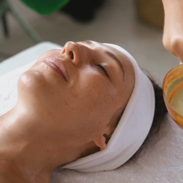 An esthetician’s hand applying French Green Clay Mask to a woman lying relaxed on a spa table, capturing a moment of skin renewal.