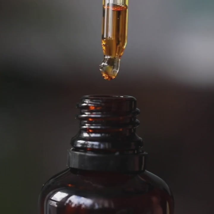 A thick golden oil dripping into an amber glass bottle, illustrating the rich texture of Coastal Winds Pomegranate Oil Eye Serum.