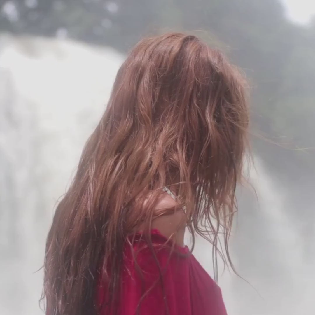 A woman with long, flowing reddish hair standing before a cascading waterfall, symbolizing the natural beauty of henna for hair.