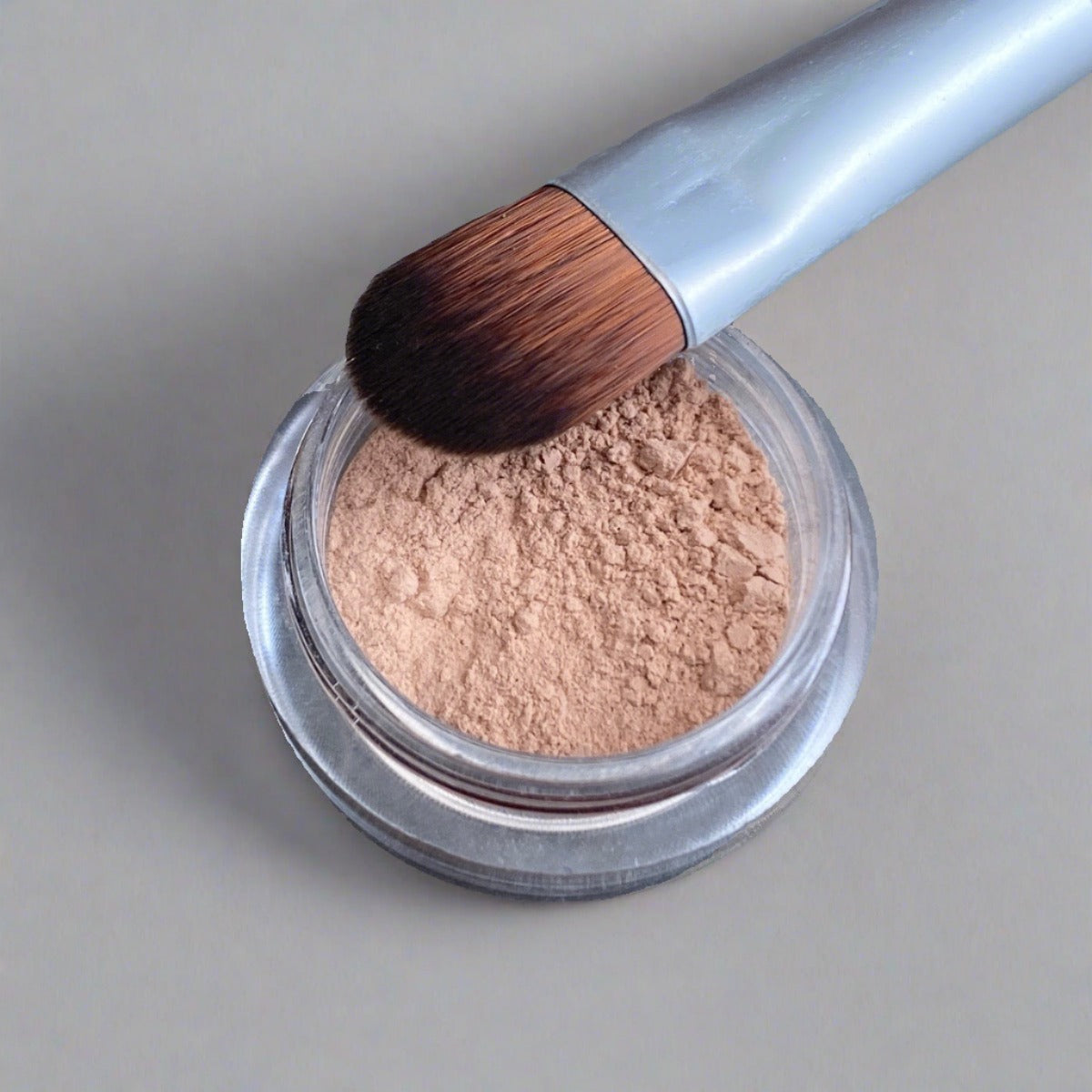 Small jar of concealer powder alongside small, dense, packing makeup brush 