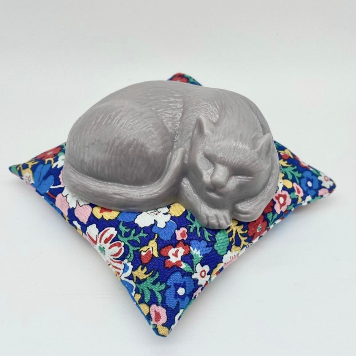 Mindfully sleeping gray cat-shaped soap, embodying the essence of mindful beauty, nestled on a vibrant floral pillow adorned with bright colors of red, blue, white, and yellow